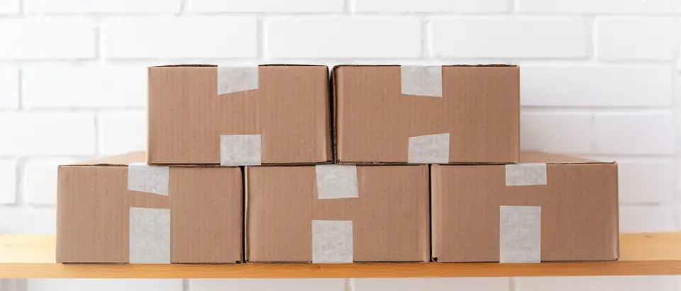 A neat stack of taped cardboard cartons on a wooden shelf against a white brick wall